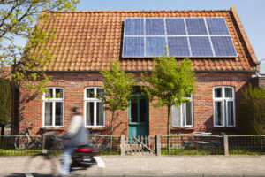 Bakstenen huis met oranje pannendak en zonnepanelen, duurzame woning met groene deur en bomen aan de voorkant, fietser in beweging op de voorgrond.