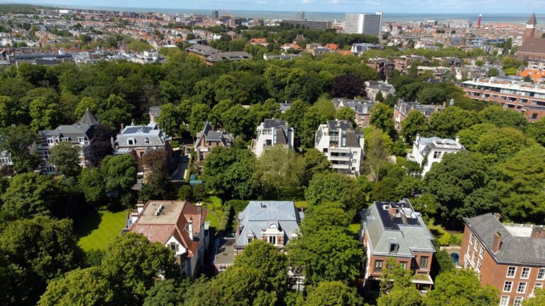 Een luchtfoto van een groen en exclusief deel van Den Haag, met indrukwekkende villa's en statige huizen omgeven door bomen, met de stad en de kustlijn op de achtergrond.