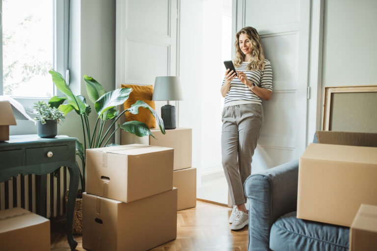 Vrouw staand in woonkamer tijdens verhuizing, omringd door verhuisdozen en planten, terwijl ze op haar telefoon kijkt.