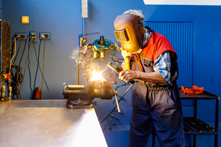 Man draagt een laskap en werkt met vonken aan een metalen object in een werkplaats, bezig met metaal bewerken